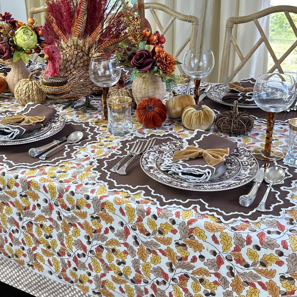 Thanksgiving table setting with fall-themed decor, including pumpkins and leaves on a patterned tablecloth.