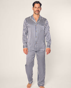 A man in a navy striped pajamas set from Petite Plume stands on a plain background. The ensemble, crafted from luxurious mulberry silk, includes a long-sleeved button-up shirt and pants. His beard and short hair add to his understated elegance.