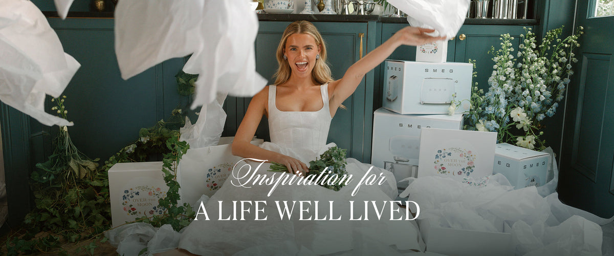 Smiling woman celebrating among gifts and flowers in a dark green kitchen with text Inspiration for a life well lived