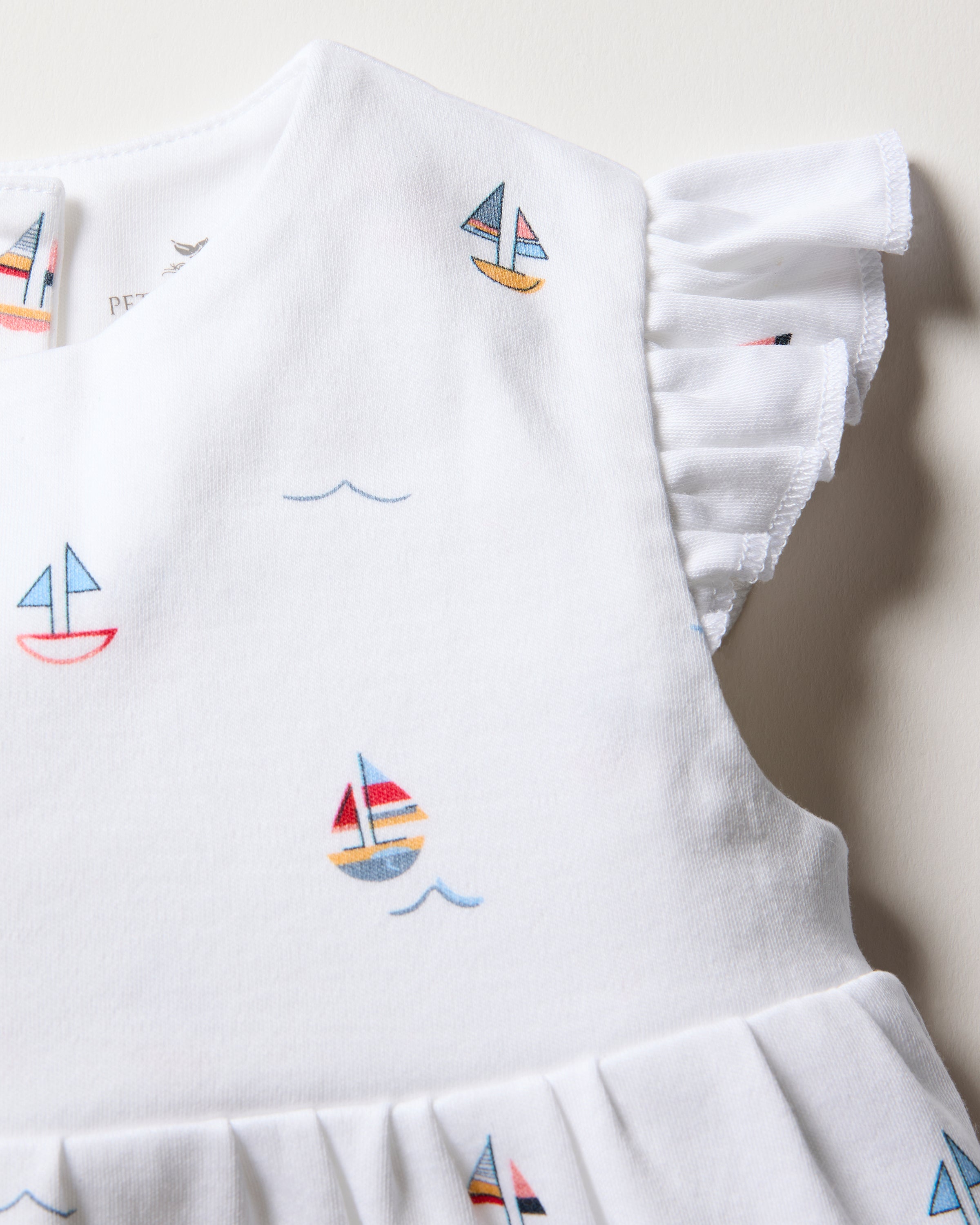 A close-up of the Babys Pima Isla Play Romper in Bateau by Petite Plume showcases vibrant sailboat and wave patterns on soft white Pima cotton, with ruffled sleeves for a playful look reminiscent of toy boats on a gentle sea.