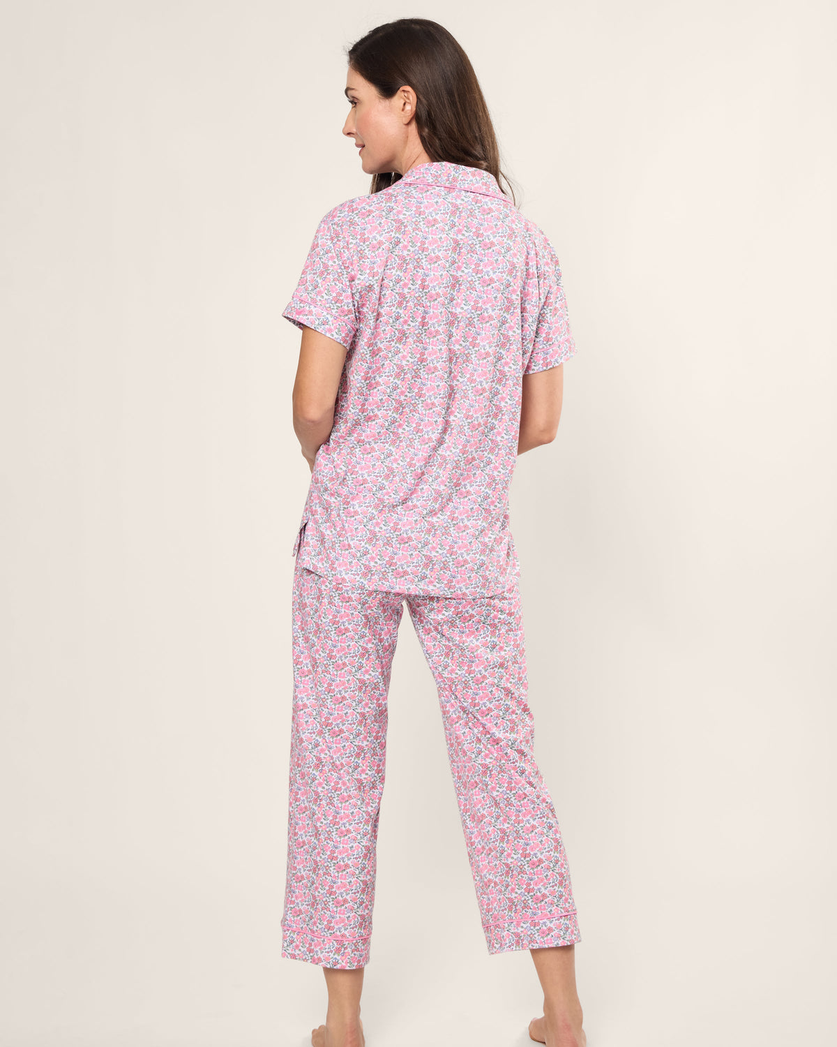 A person stands barefoot facing away in a Womens Pima Crop Set in Fleurs de Rose by Petite Plume, featuring a pink and white floral pattern with soft Pima cotton short sleeves and long pants, set against a plain, light-colored background—ideal for any bedtime ritual.