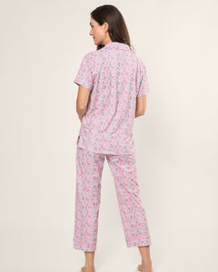 A person stands barefoot facing away in a Womens Pima Crop Set in Fleurs de Rose by Petite Plume, featuring a pink and white floral pattern with soft Pima cotton short sleeves and long pants, set against a plain, light-colored background—ideal for any bedtime ritual.