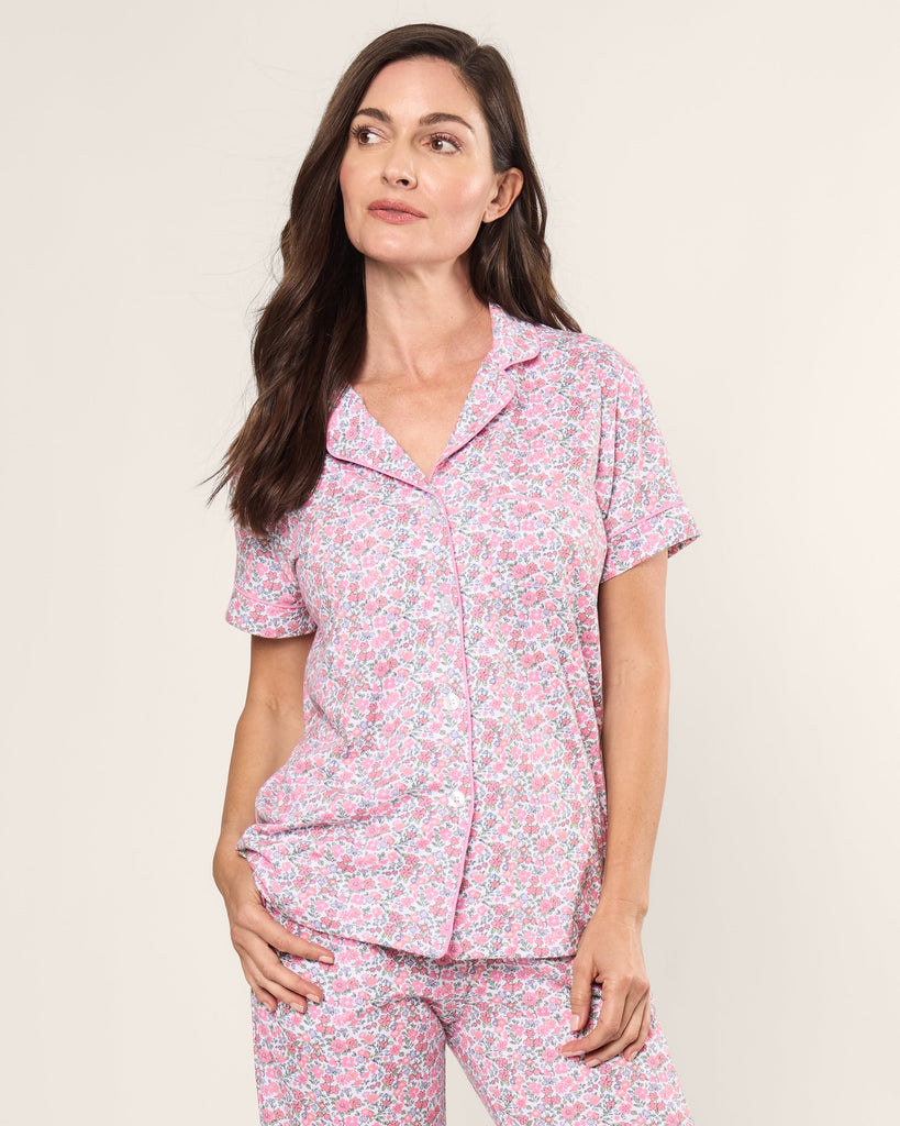 A woman with long dark hair wears the Womens Pima Crop Set in Fleurs de Rose by Petite Plume, featuring short sleeves and a pink floral design in soft Pima cotton, against a plain backdrop. She looks to the side with a relaxed expression, encapsulating bedtime serenity.