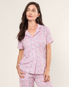 A woman with long dark hair wears the Womens Pima Crop Set in Fleurs de Rose by Petite Plume, featuring short sleeves and a pink floral design in soft Pima cotton, against a plain backdrop. She looks to the side with a relaxed expression, encapsulating bedtime serenity.