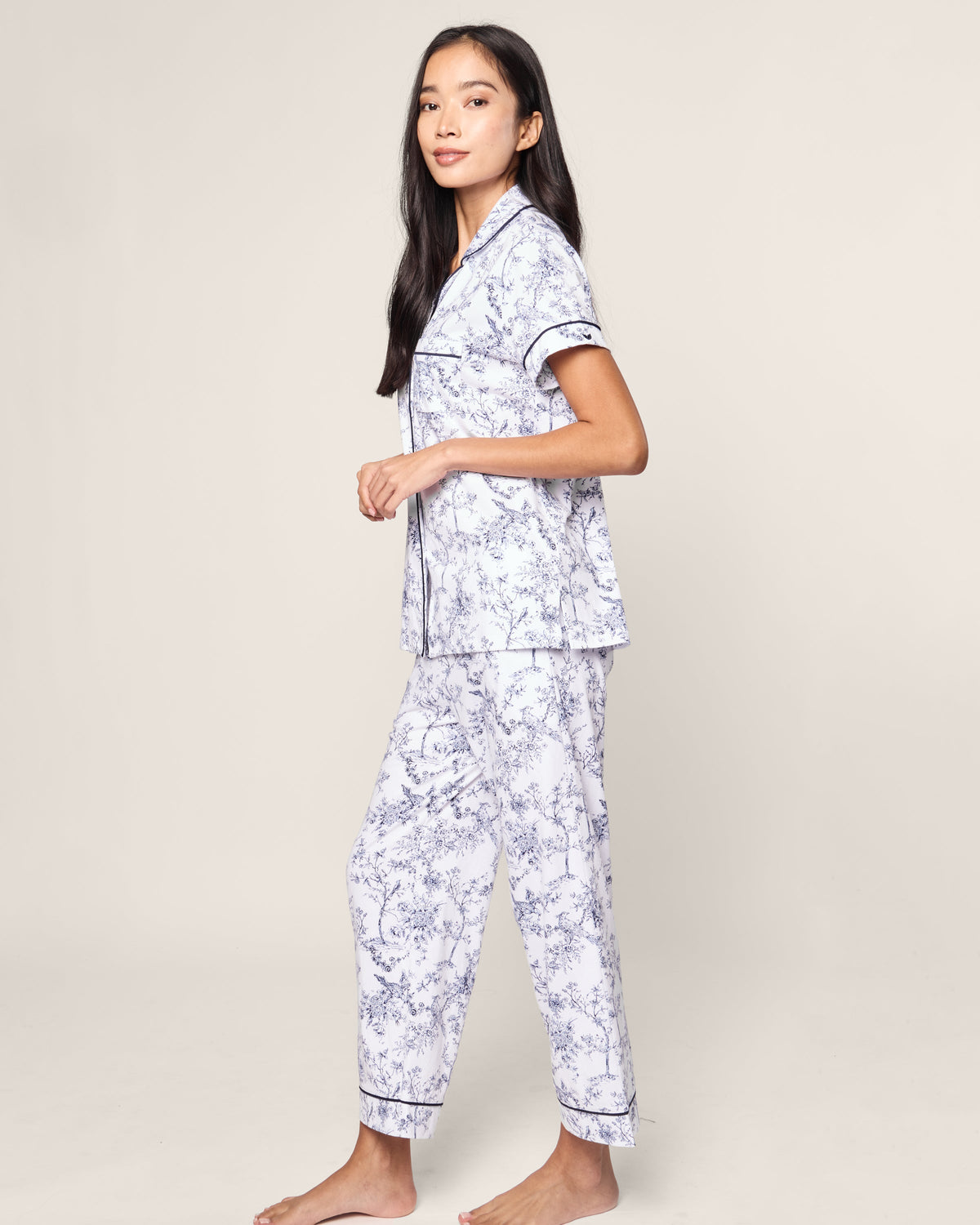 A woman stands sideways in the Womens Pima Crop Set in Timeless Toile by Petite Plume, featuring short sleeves and a button-up design with classic floral patterns, against a neutral background.