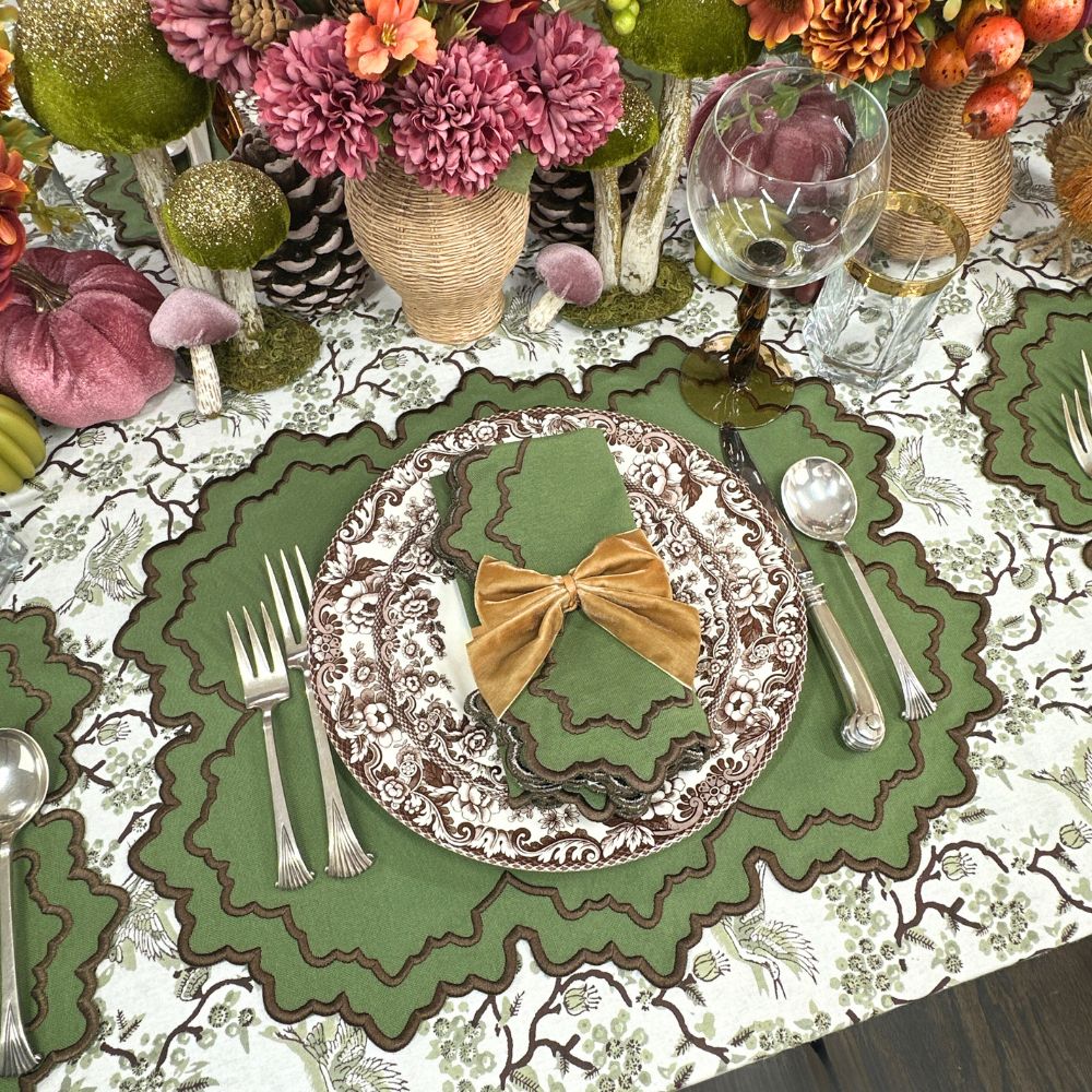 Decorative table setting with a plate featuring a bow, surrounded by cutlery and place mats on a patterned toile sage tablecloth.