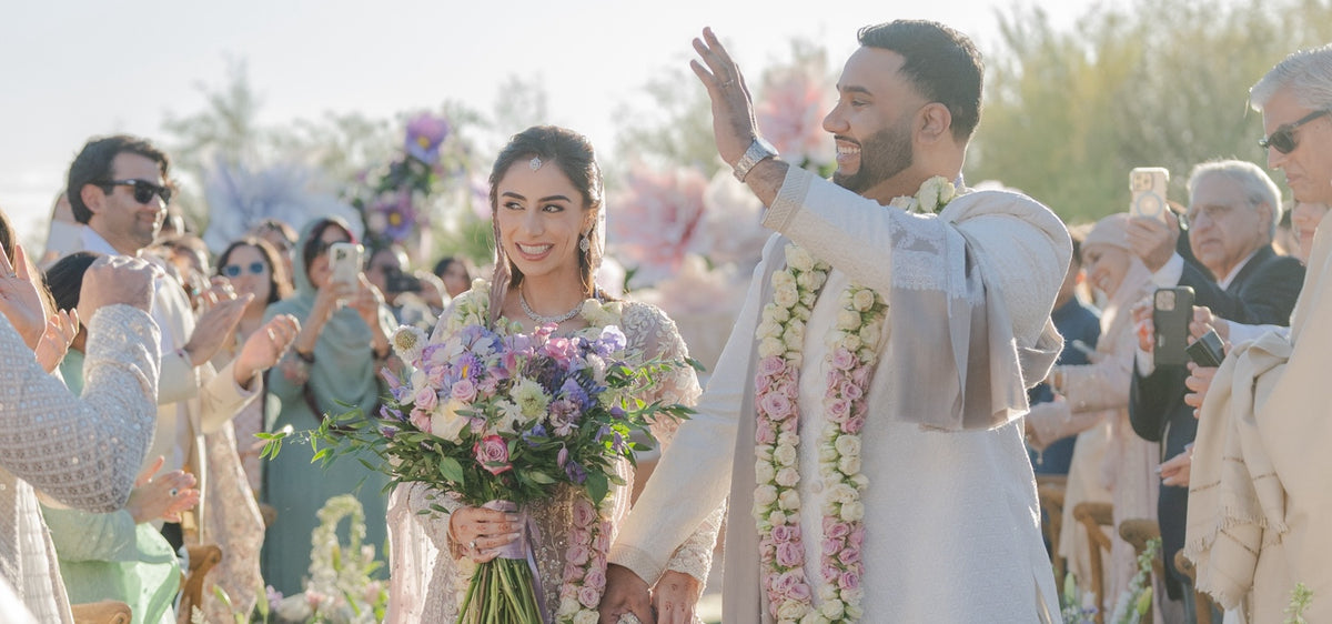 Newlywed couple walk through an outdoor wedding ceremony as guests take photos, with bouquet and flower garlands