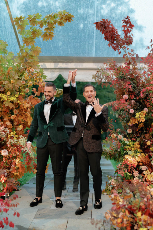 Three men in formalwear celebrate at an autumn wedding surrounded by colorful flowers and foliage