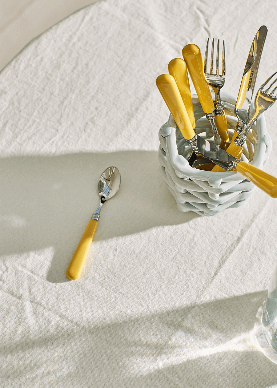Angled view of small sky blue basketweave planter showing woven texture with yellow handled flatware on linen tablecloth