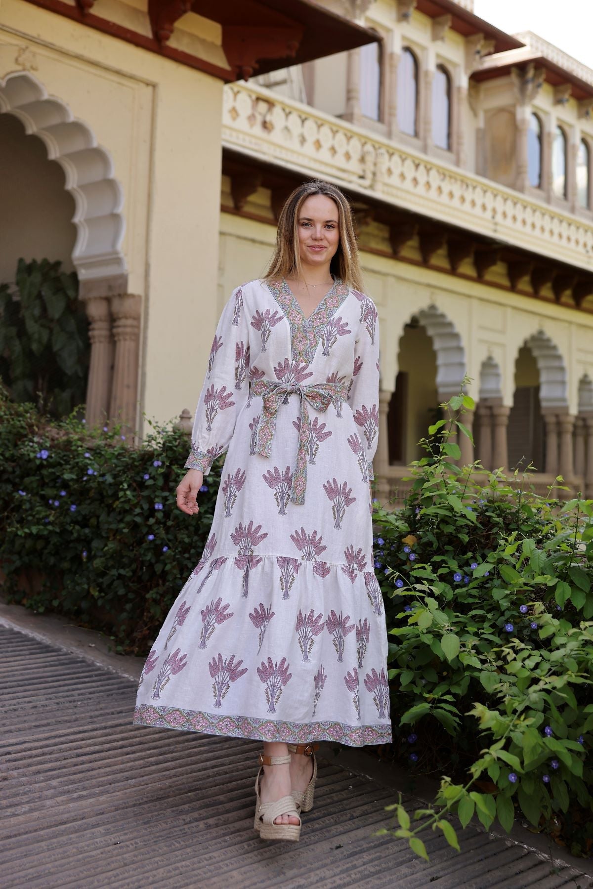 Front view of Capri maxi dress in Pink Palm showing V-neck, long sleeves, self-tie waist and tiered skirt against arched courtyard and greenery