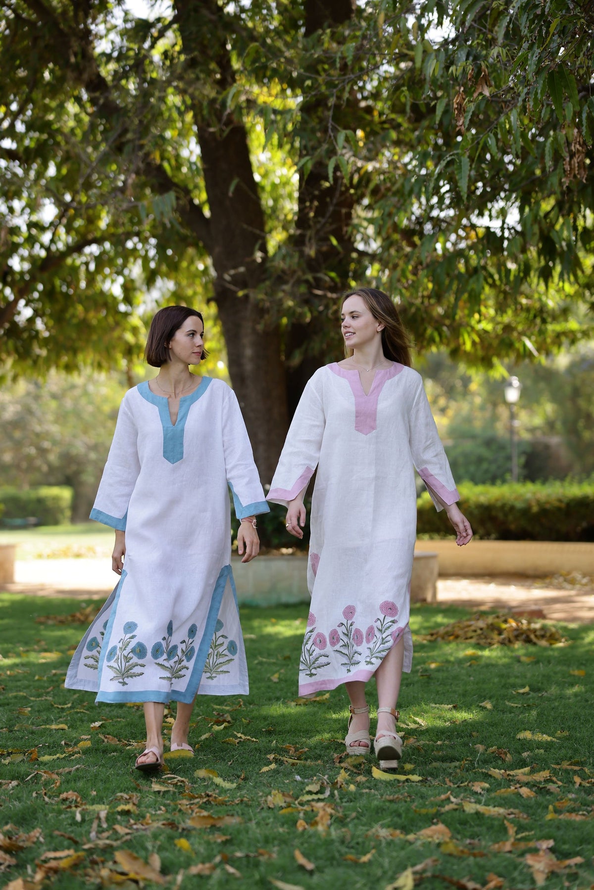 Two models wearing Hampton Caftans, one pink-trimmed and one blue-trimmed, showing embroidered floral hem while walking in a park