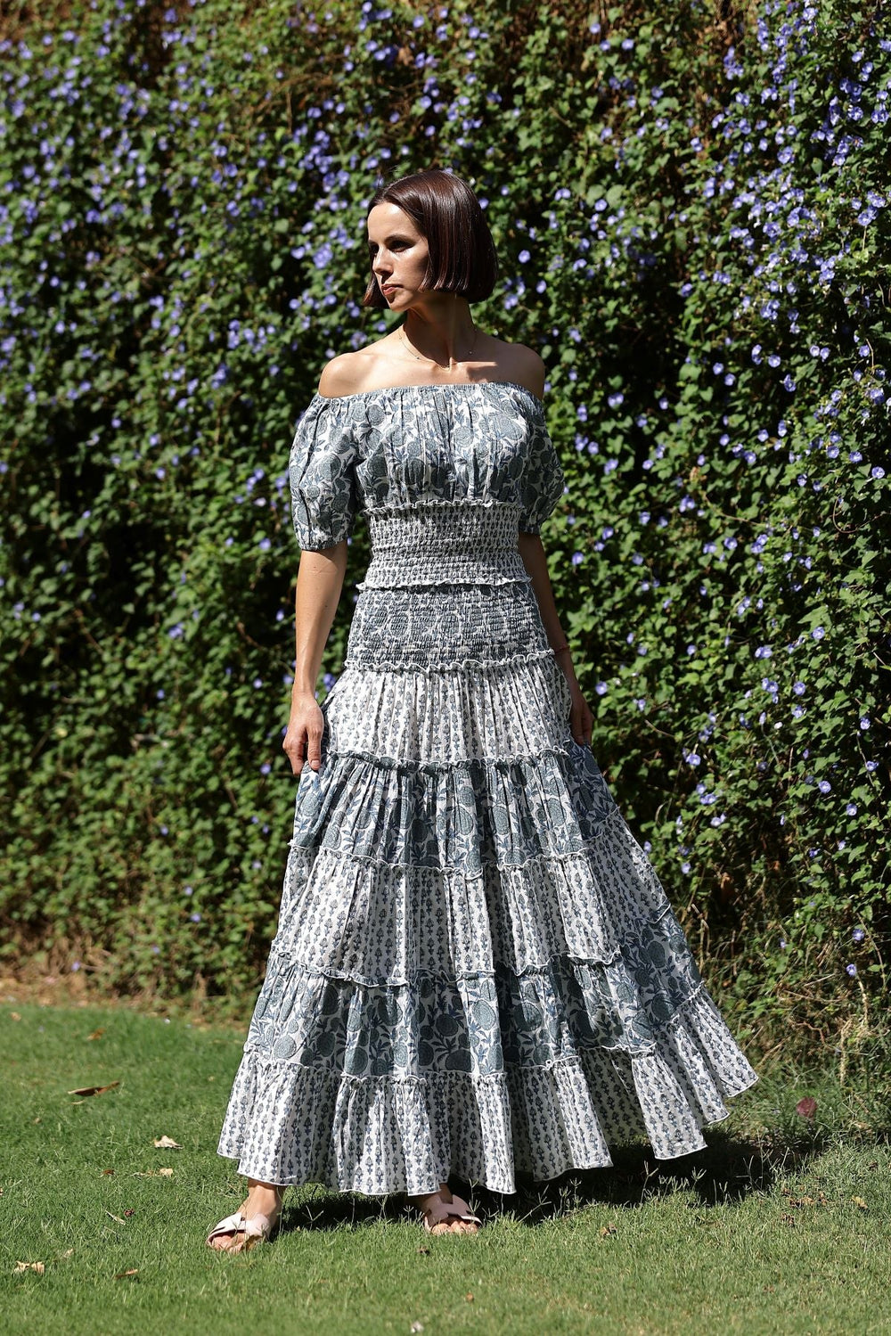 Three-quarter view of Bora Bora skirt in Blue Marigold with smocked drop waist, tiered ruffles and hidden side pockets against a green garden hedge