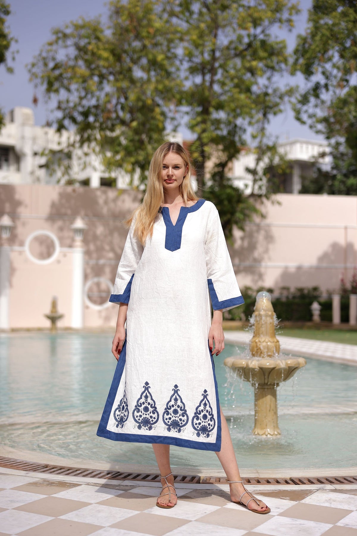 Front view of Hampton Caftan showcasing blue-trimmed V-neck, 3/4 sleeves, and indigo paisley embroidered hem with model standing by a fountain