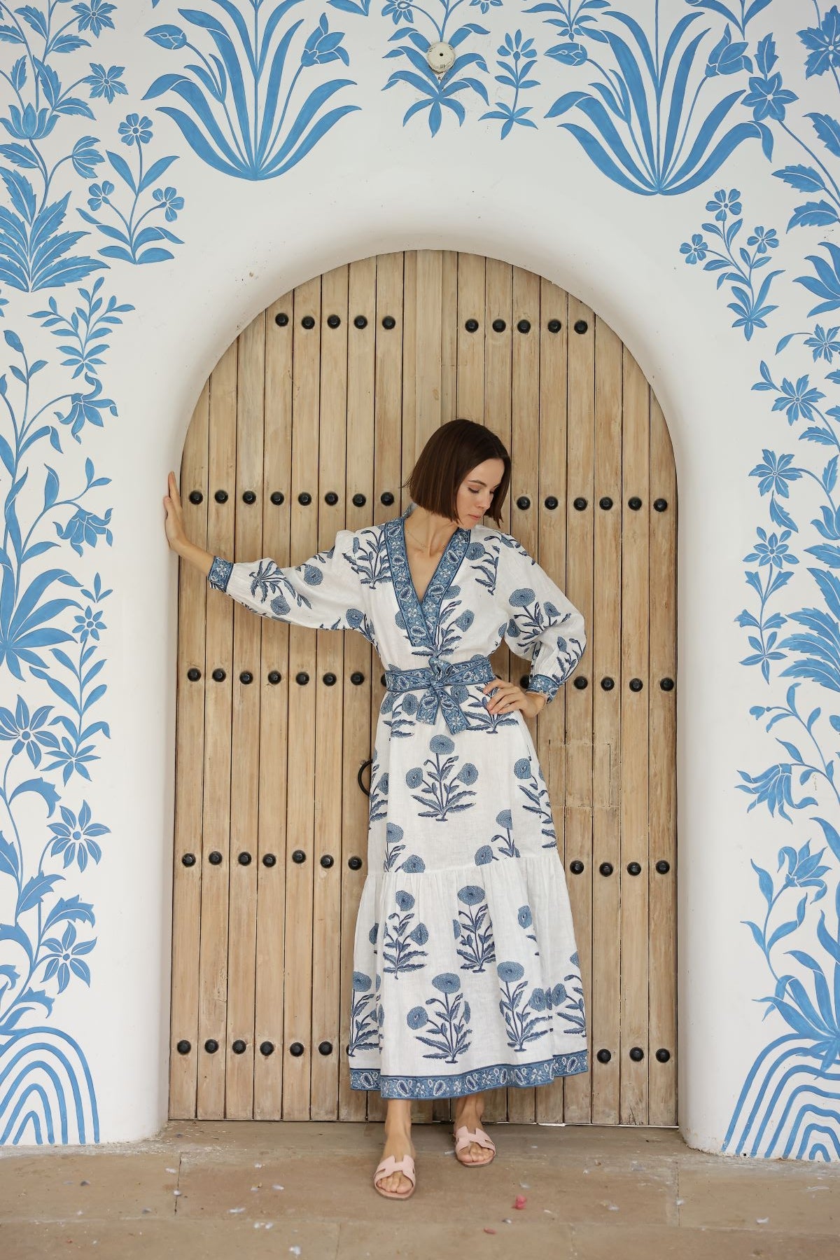 Front view of Capri Maxi Dress in indigo with V-neck, sleeves, tie waist, blue floral print on painted archway background