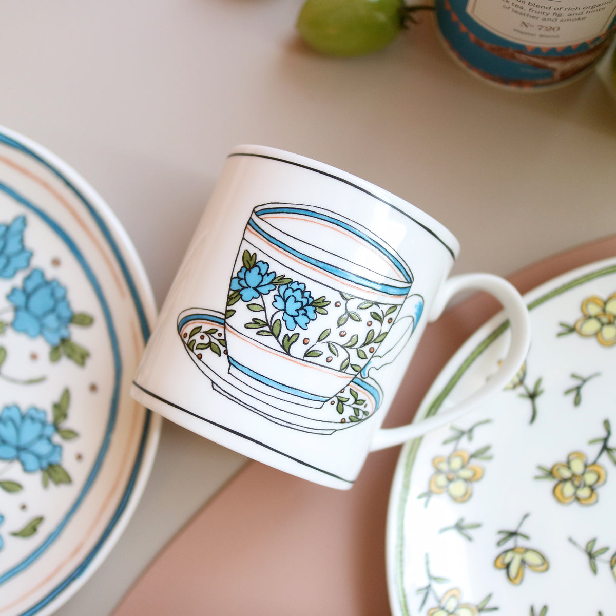 Angled view of Heritage Bachelor Button Mug showcasing blue cornflower decals with floral china in the background