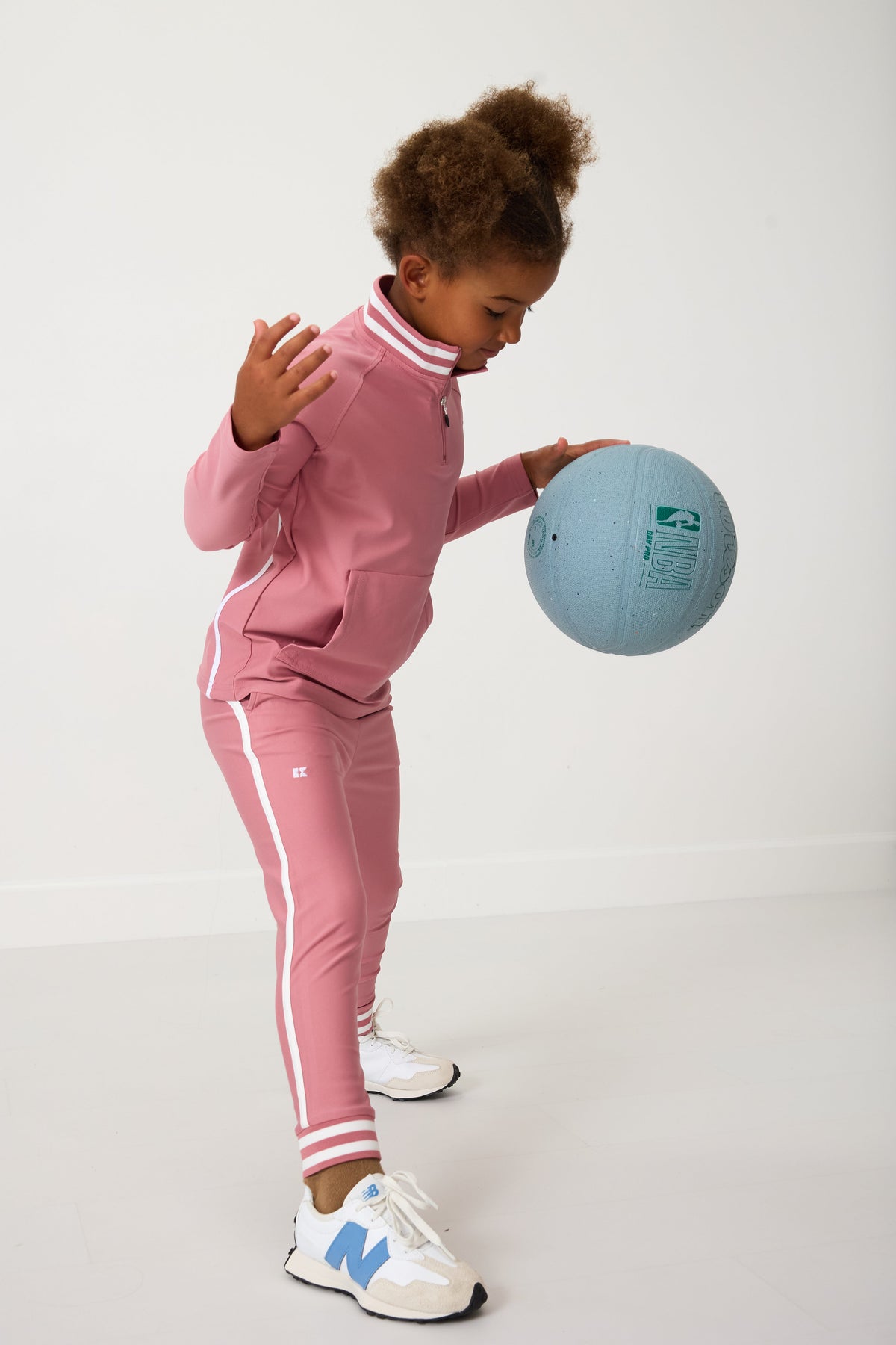 Three-quarter view of Warm-Up Jogger in Dusty Pink on child model showcasing slim fit, white side stripes, and above-ankle length in plain studio