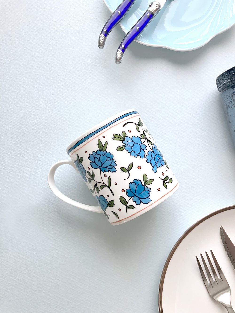 Angled view of Heritage Bachelor Button mug showcasing blue floral pattern on white fine bone china against blue tabletop
