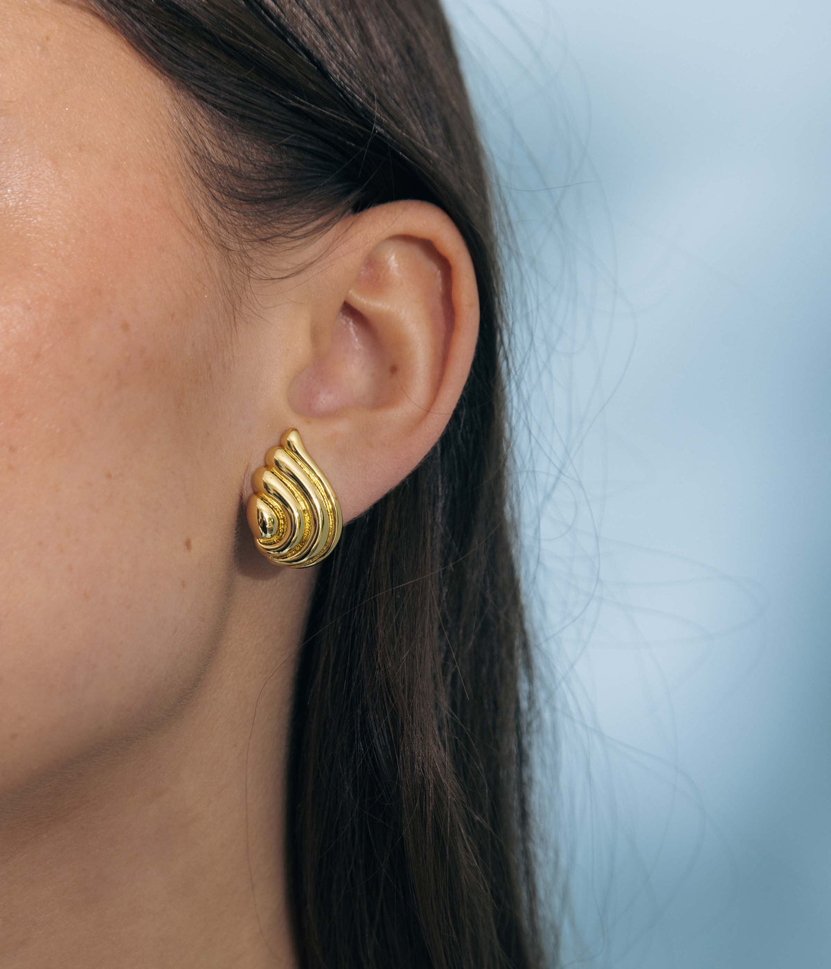 Close-up of Florina Stud Earrings in Gold showcasing sculptural winged silhouette on a model against pale blue background