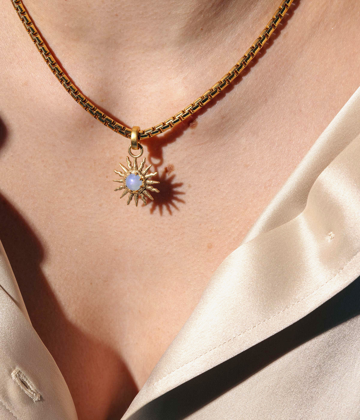 Close-up of the Fira Sun Pendant in Gold showcasing its hammered sunburst design and pale blue cabochon on beige fabric