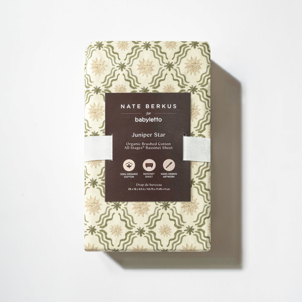 Top view of All Stages Bassinet Sheet in Juniper Star showcasing folded patterned fabric and product label on white background