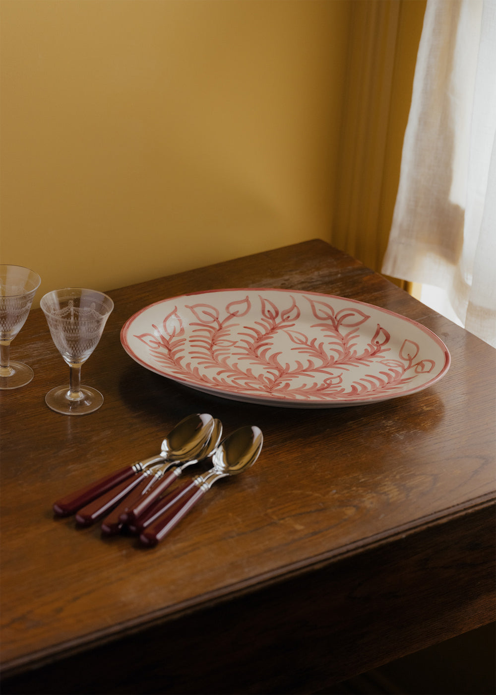 Large pink Vina platter with hand-painted leaf motif on a wooden table beside spoons and glassware