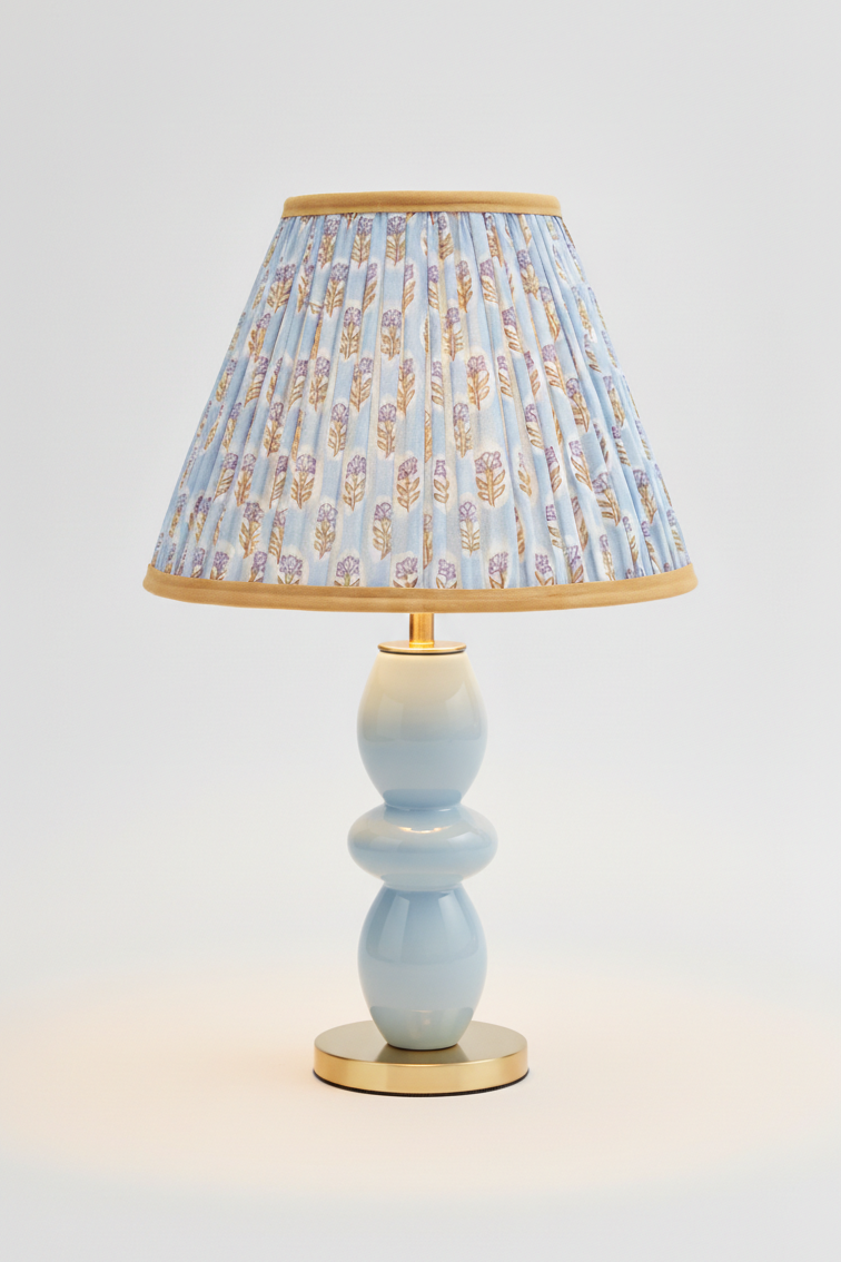 10-inch pleated blockprint empire lampshade with sky blue daisy pattern, mounted on light blue ceramic table lamp with golden base, illuminated with warm light