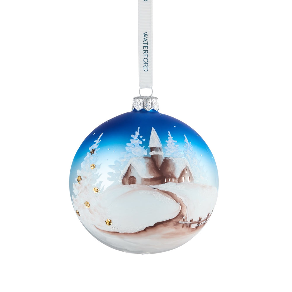 Front view of Holiday Heirlooms Winter Scene glass ornament showing hand-painted snowy village, blue ombré sky, gold sequin accents, and white Waterford ribbon