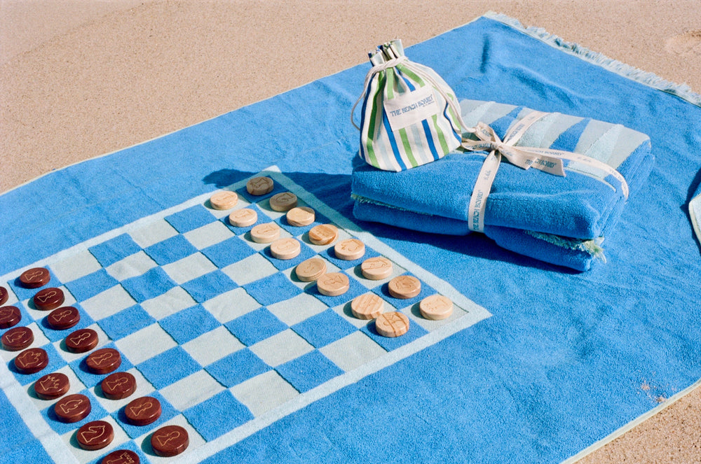 Angled view of The Montauk Beach Board towel showcasing regulation-size chess and checkers print with wooden game pieces and canvas pouch on sand