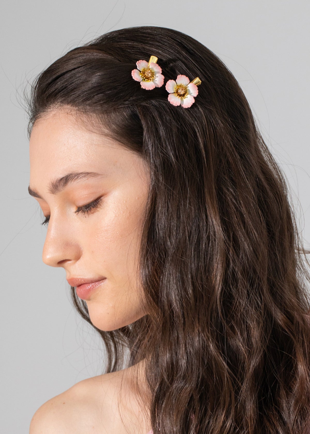 Side view of Debbie hair clips in blush showing two pink flower clips with gold centers worn in a model’s dark hair on gray