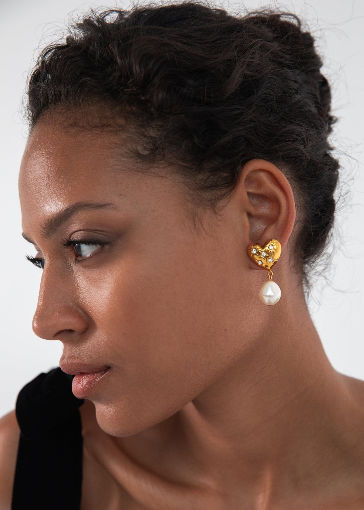 Side view of Hayden Earrings in Crystal Antique Gold showcasing a crystal-studded heart and dangling baroque pearl on model