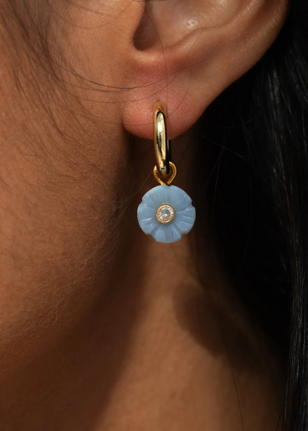 Close-up of Lillian earring in blue opal showing hand-carved floral charm with center crystal on a gold-plated huggie worn on a model’s ear