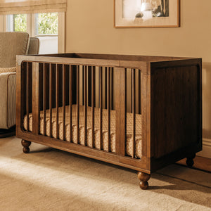 Front angled view of the Toffee Astre All Stages 8 in 1 crib showcasing vertical slats and caster wheels in a warm nursery