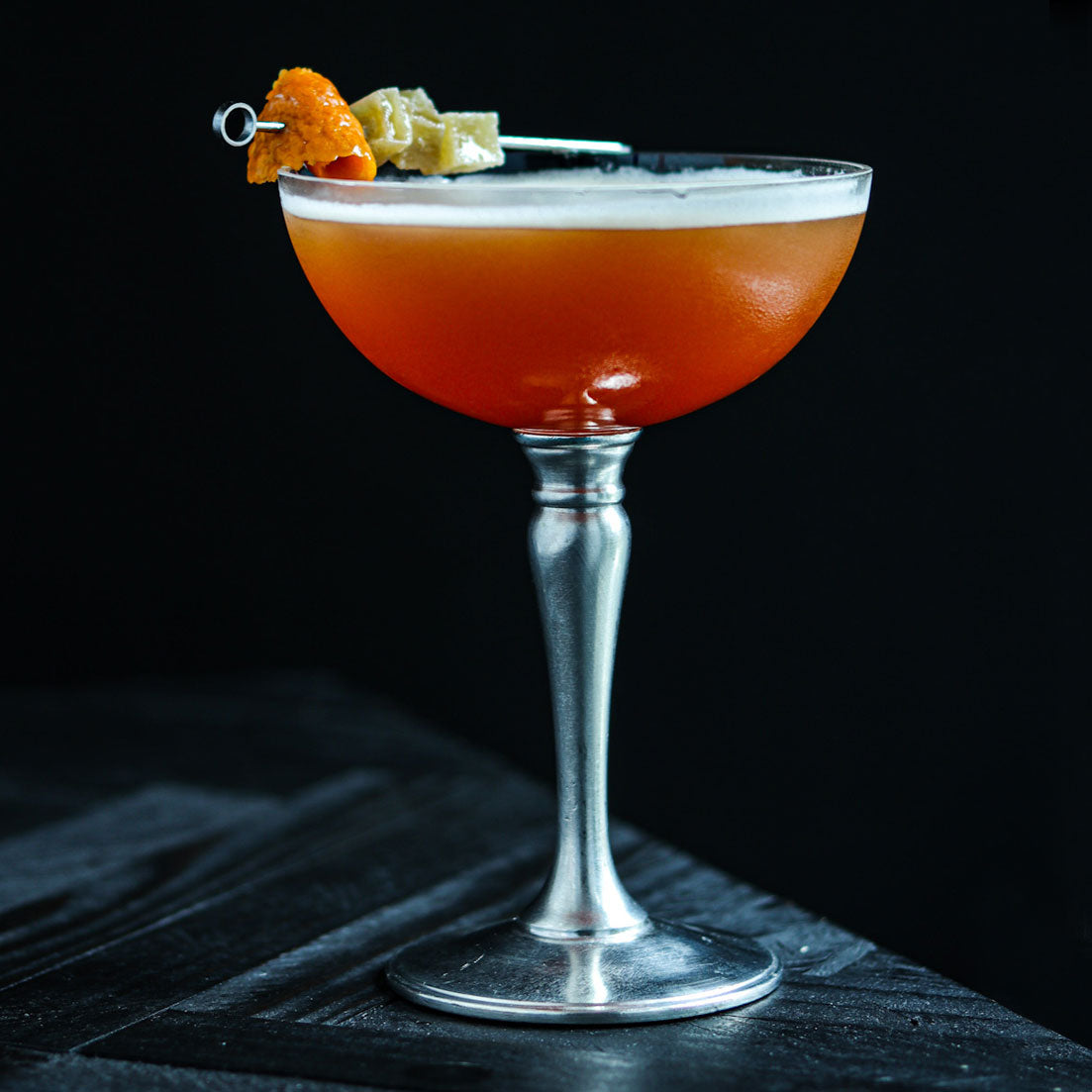 Side view of Champagne and Cocktail Coupe with orange cocktail and foam, pewter stem and base, orange peel garnish