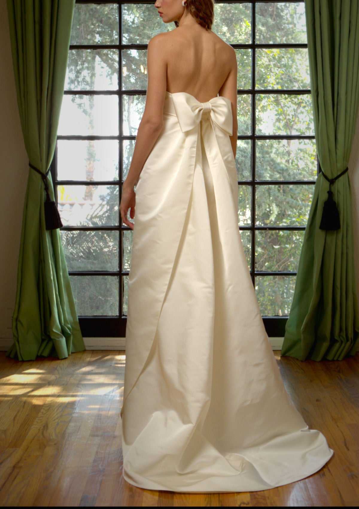 Back view of Strapless Petal Gown in creme peau de soie showcasing low back, large bow and floor-length train before a paned window
