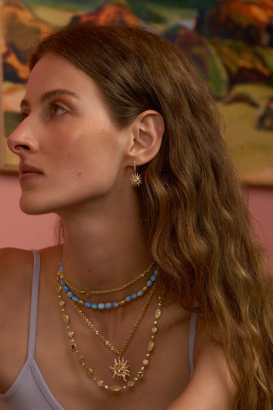 Left profile of model wearing El Sol earrings showcasing dangling sun motif in 14K yellow gold with diamond accents against a pink background