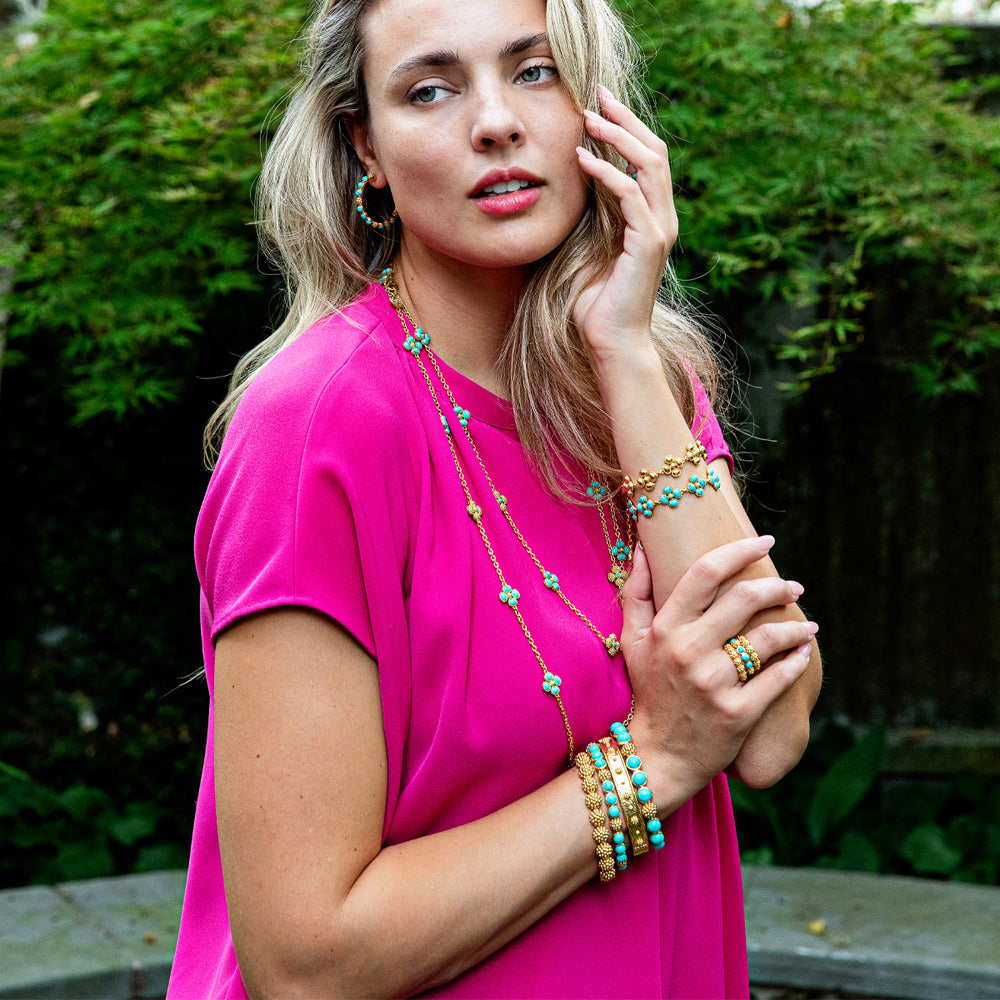 Model wearing a turquoise berry gem hinged bangle, stacked turquoise and gold bracelets, and matching necklace in an outdoor garden setting