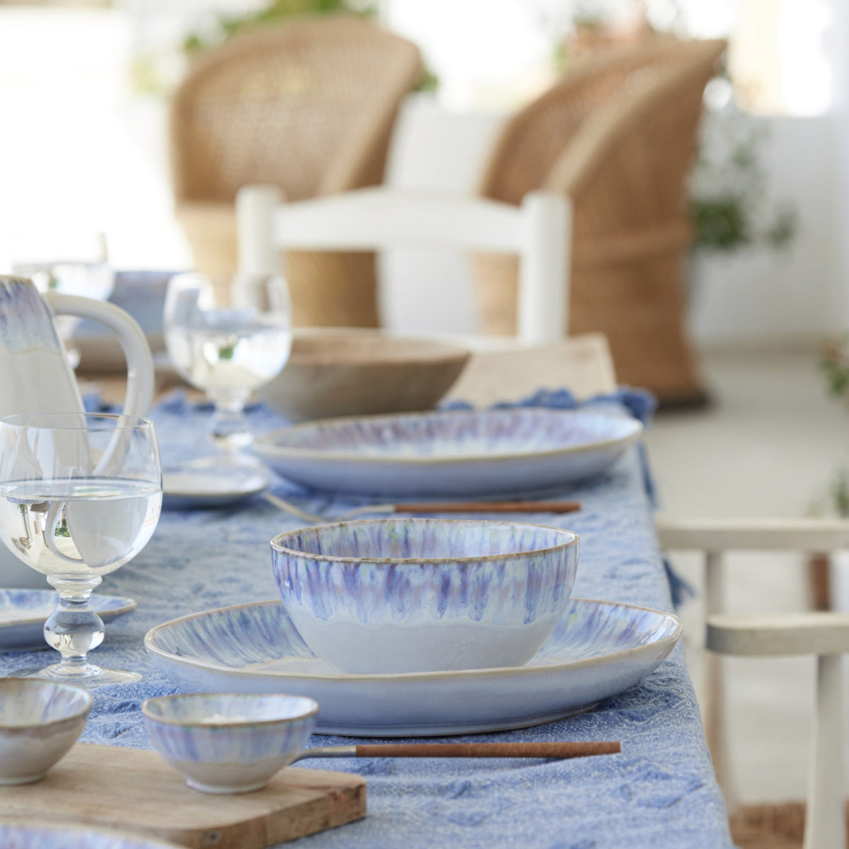 Brisa cereal bowls arranged on a blue linen table with matching plates and glassware in a sunlit coastal setting