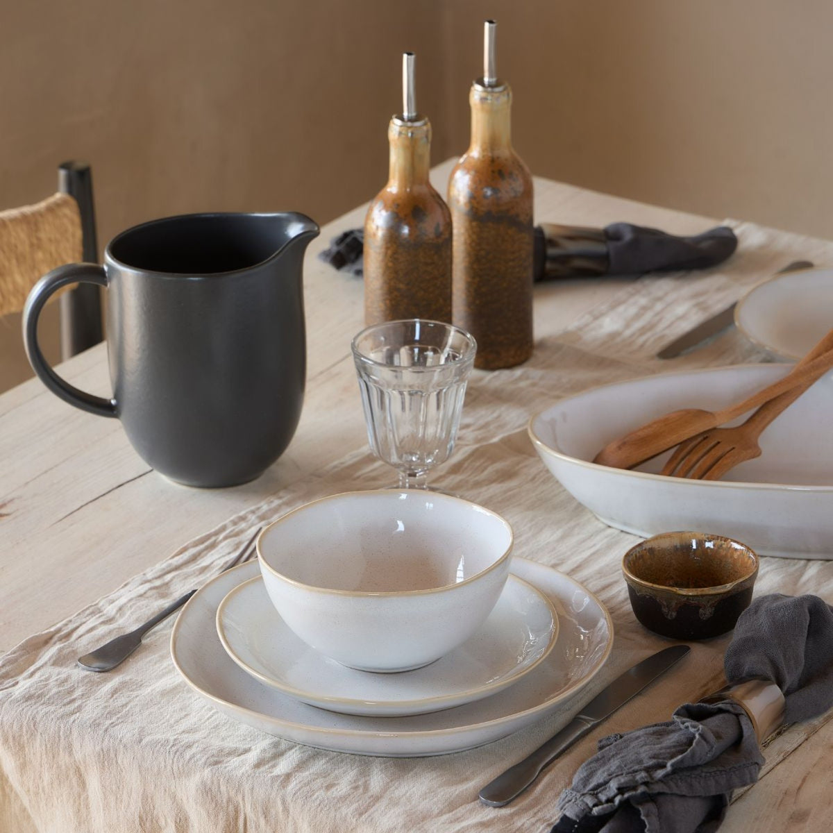 Brisa Collection Branca oval baker on a linen-covered dining table in a styled rustic place setting with matching white bowls and a dark pitcher