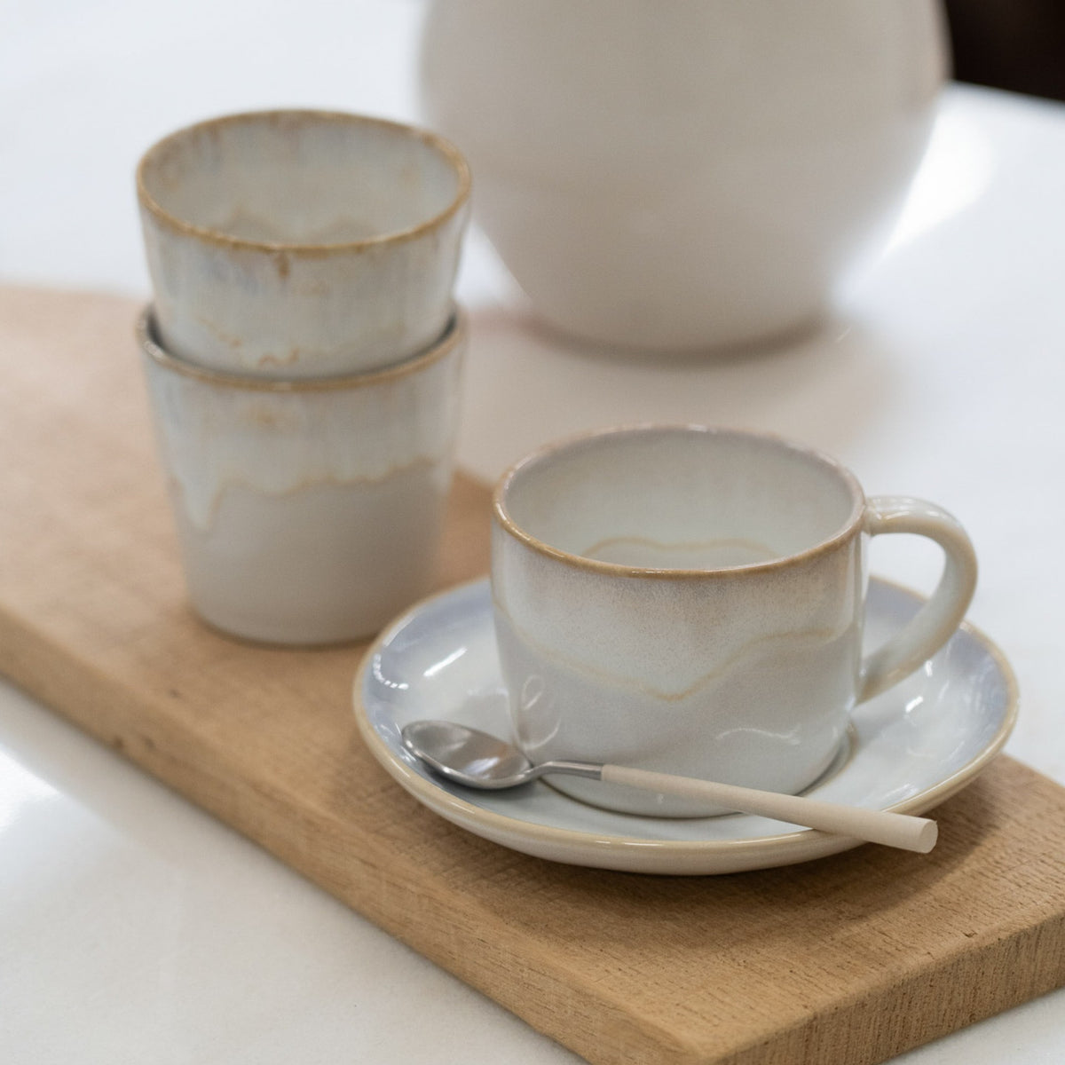 Front view of Brisa cup and saucer in salt glaze with gold rim on a wooden board, stacked cups and teaspoon