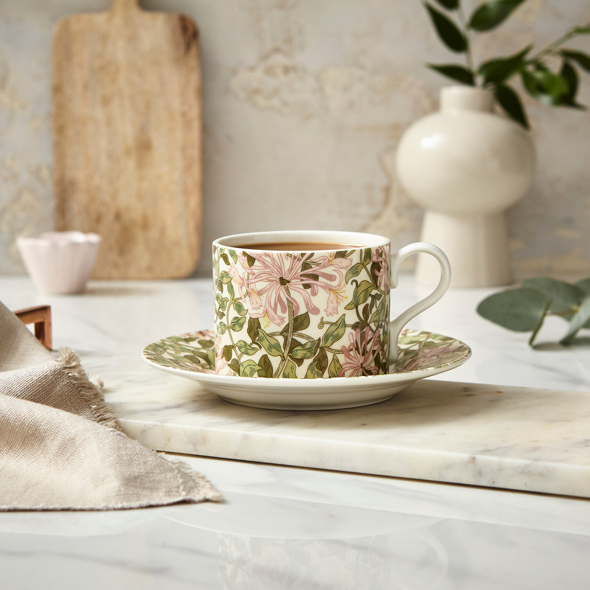 Front view of Morris and Co. Teacup and Saucer in Honeysuckle showcasing pink flowers and intertwining leaves on a light tabletop