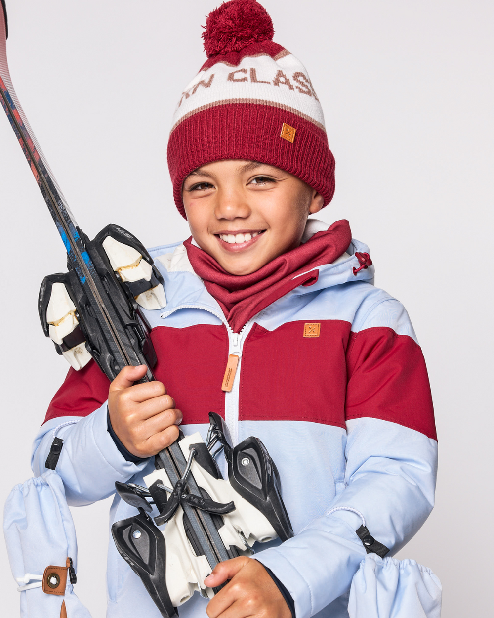 Child wearing a heritage red fleece-lined neck warmer, smiling while holding skis against a light studio background