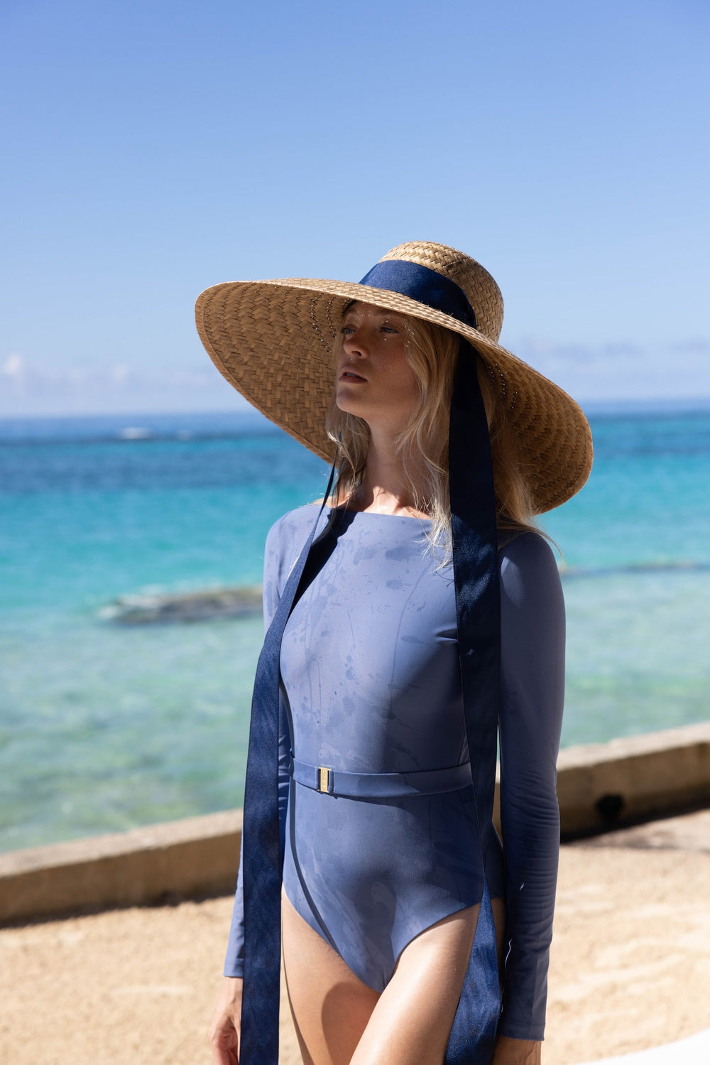 Three-quarter view of Geranium Sun Hat on model, natural woven straw with extra-wide brim and long navy ribbon ties at the beach