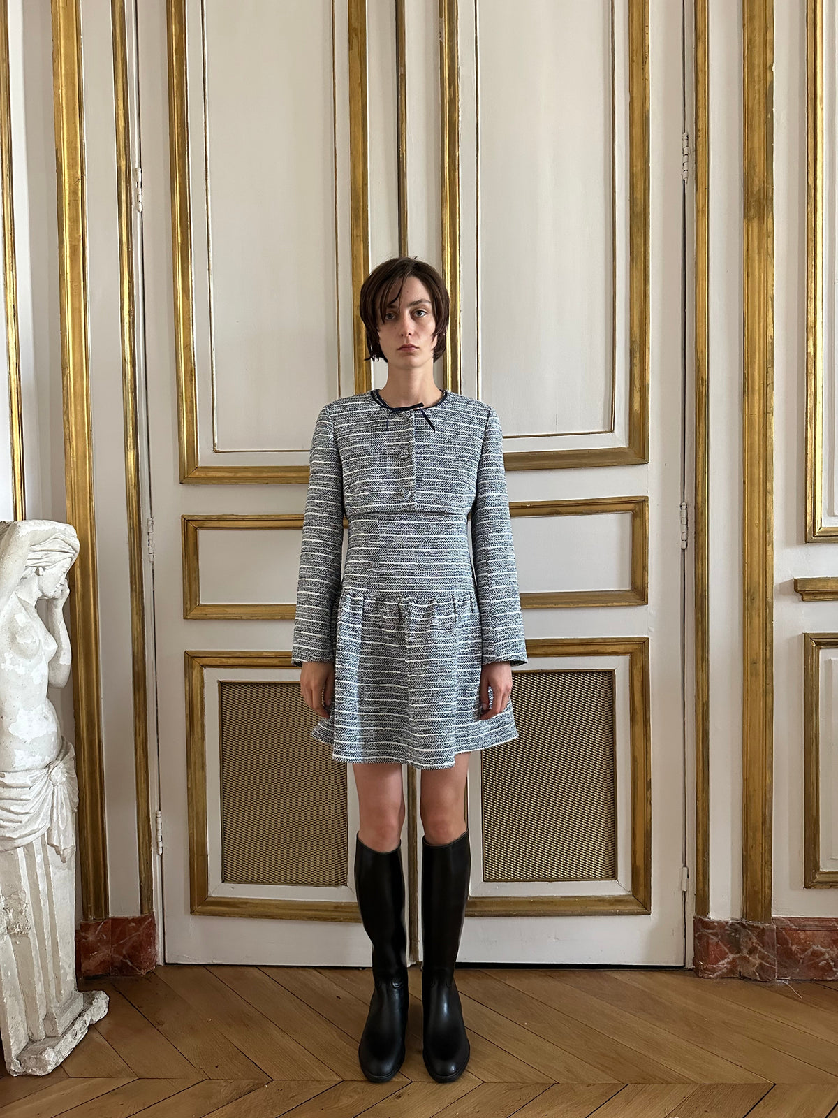 Front view of Leandra Jacket in Bon Voyage tweed with grosgrain neck bow and tweed-covered buttons on a gold-trimmed door