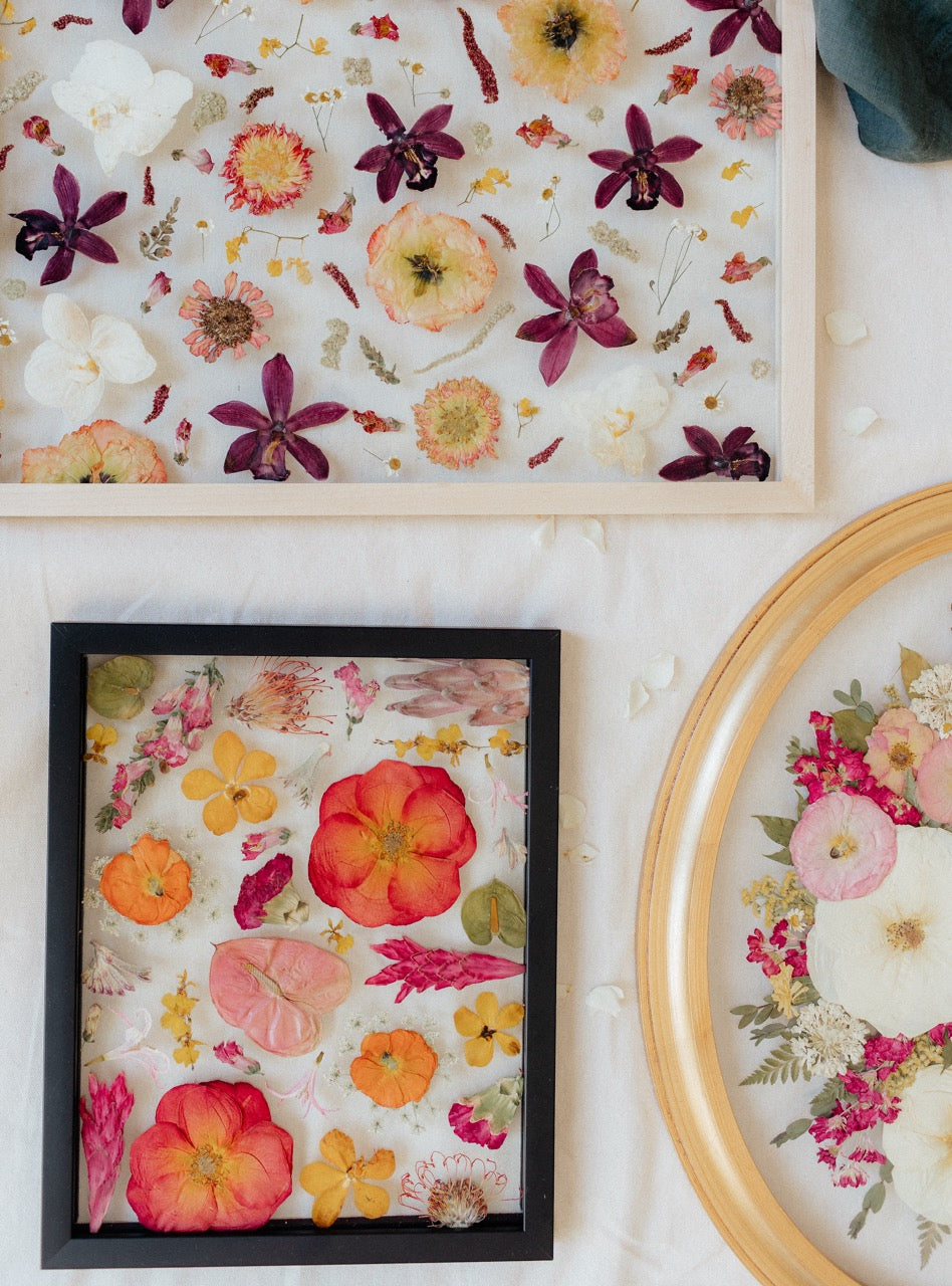 Flat lay of three pressed floral frames showcasing colorful dried flowers in wood and black frames on cream fabric background