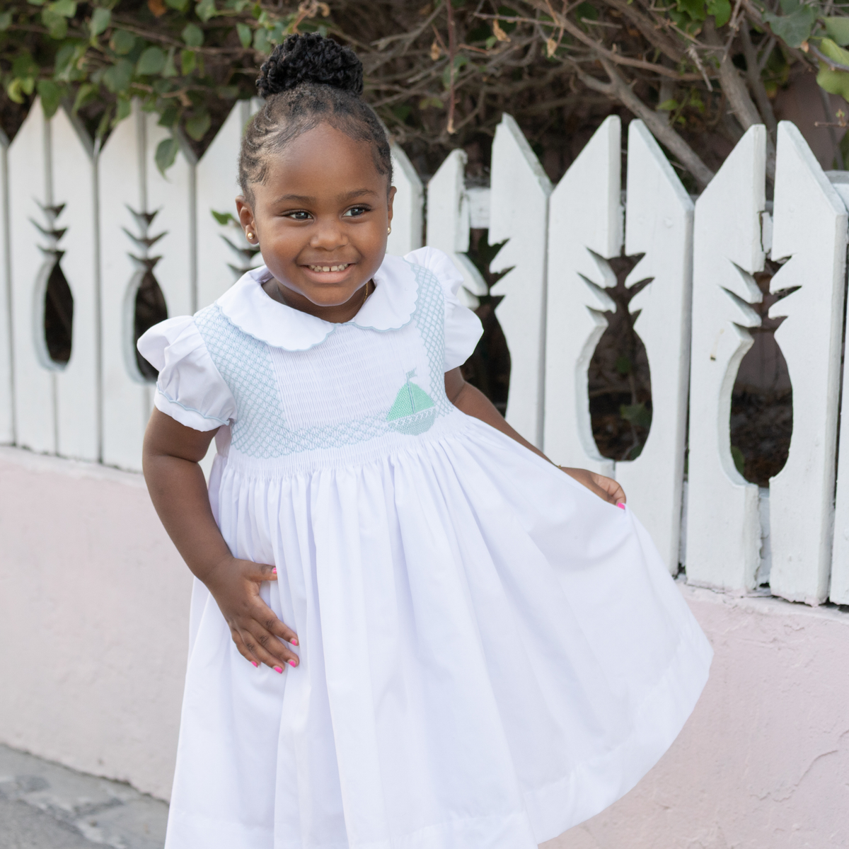 Front view of Eloise dress in white cotton showing blue smocked sailboat detail and scalloped Peter Pan collar on toddler by white picket fence