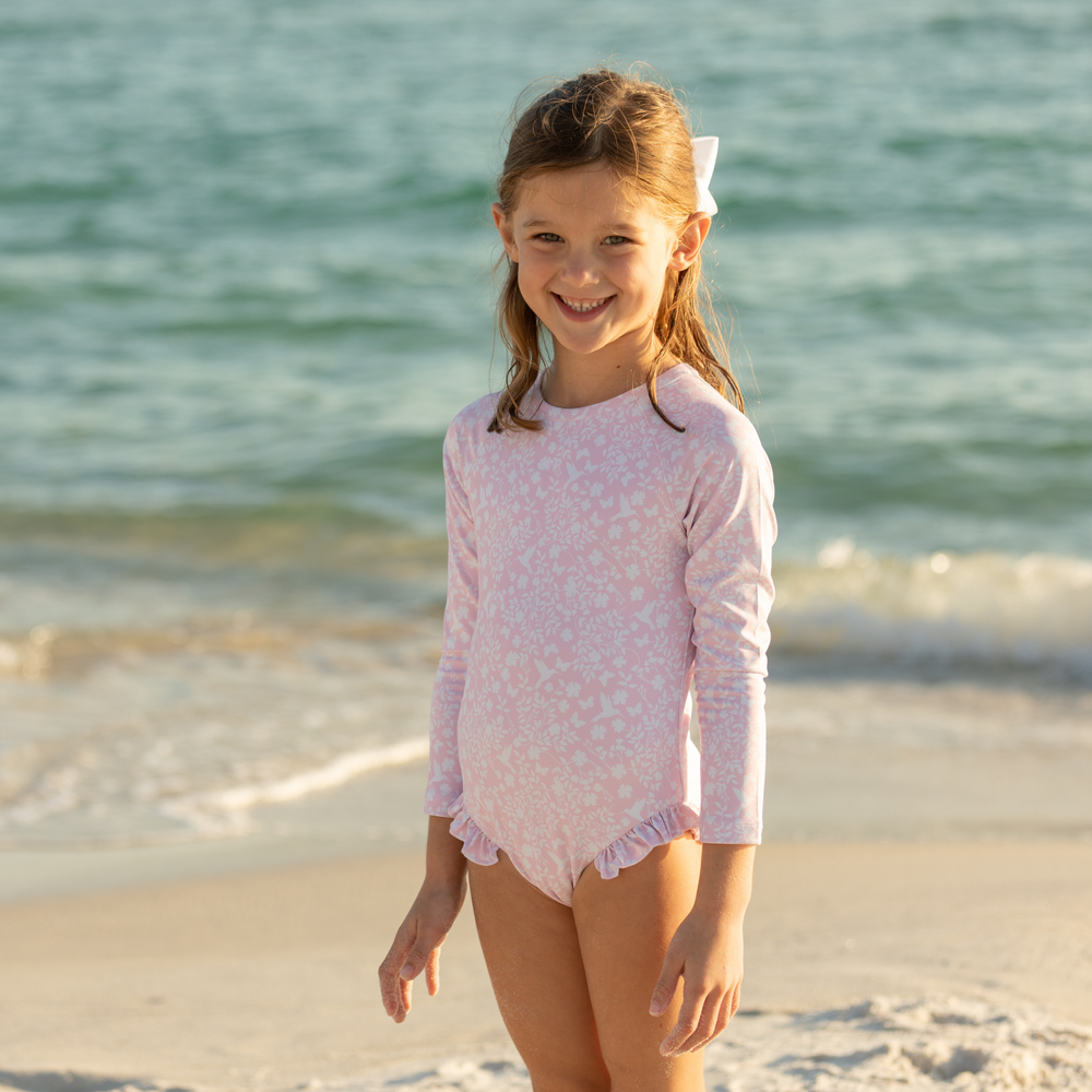 Front view of Reese Rashguard one-piece in Coral Sand Chinz, long sleeves, high neckline, ruffled leg trim, on girl at beach