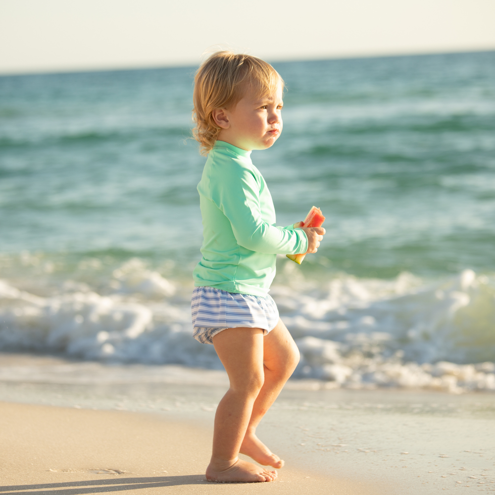 Toddler standing at the shoreline holding a slice of watermelon, wearing Bay Street Stripe swim shorties and a mint long-sleeve rash guard