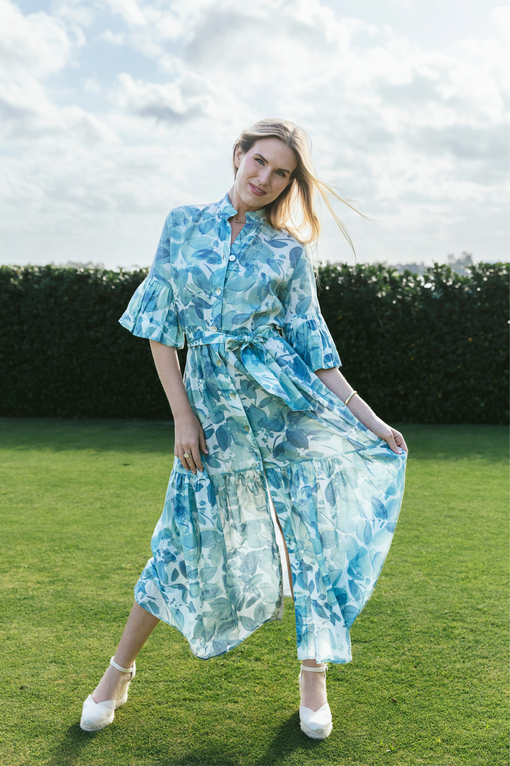 Front view of Joanna dress in blue watercolor floral linen showcasing bell sleeves, button-front bodice, tied sash, and flounced skirt on a grassy lawn