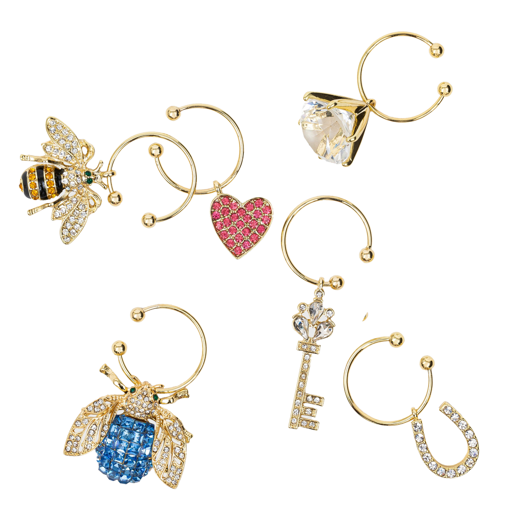 Top-down view of six gold-tone wedding wine charms showcasing bee, blue jeweled bee, pink heart, large faceted gem, key, and horseshoe on white background