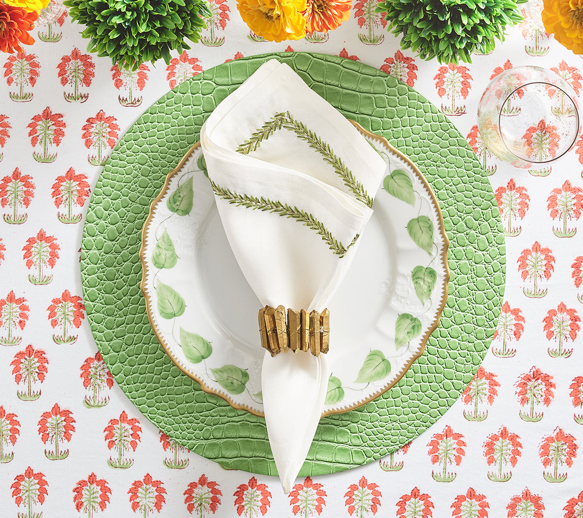 Top view of Jardin napkin folded on a plate showing green embroidered vine border and gold napkin ring on textured charger